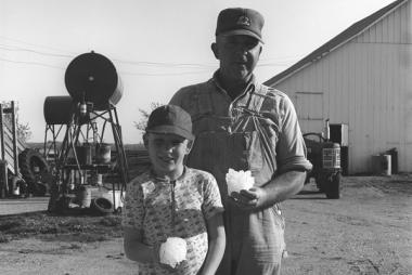 A farmer and his son, residents of rural Missouri, stand outside. Each hold a large hailstone. There is farm equipment in the background.